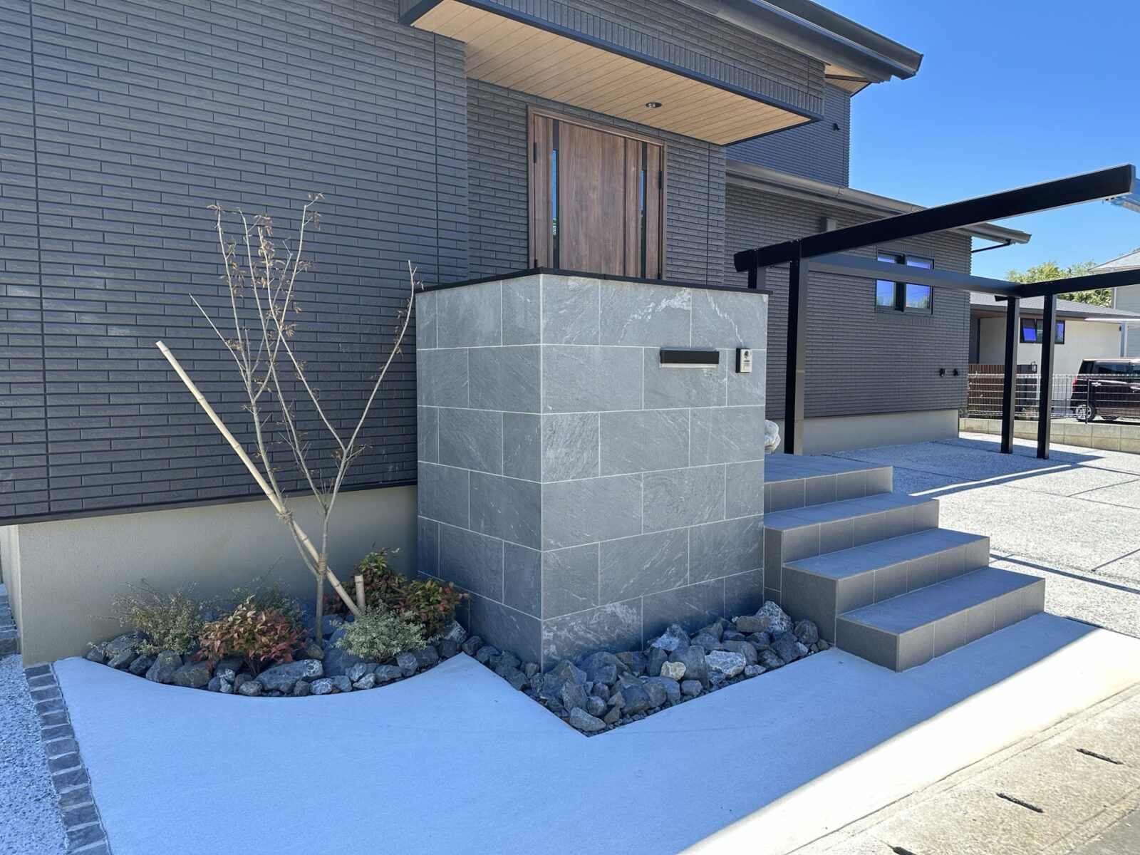 ダークグレーのモダンな外壁が特徴の住宅外観。石調タイルの門柱にはポストとインターホンが設置され、木目調の玄関ドアへ続く3段の階段とコンクリートのアプローチが印象的。足元には岩と低木の植栽が配され、スタイリッシュなエクステリアデザインが際立つ施工事例。