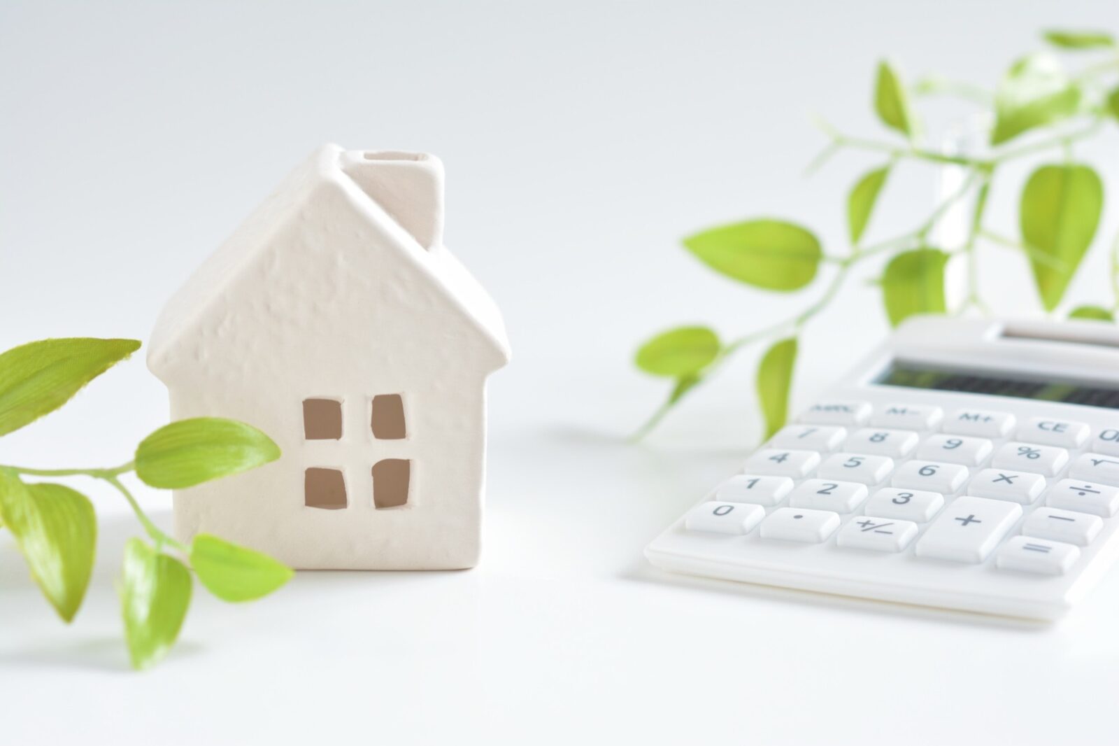 白い電卓と、白のミニチュアハウスがテーブルに並べられている。住宅ローン計算、マイホーム購入、不動産投資、家計の資金計画をイメージさせる。