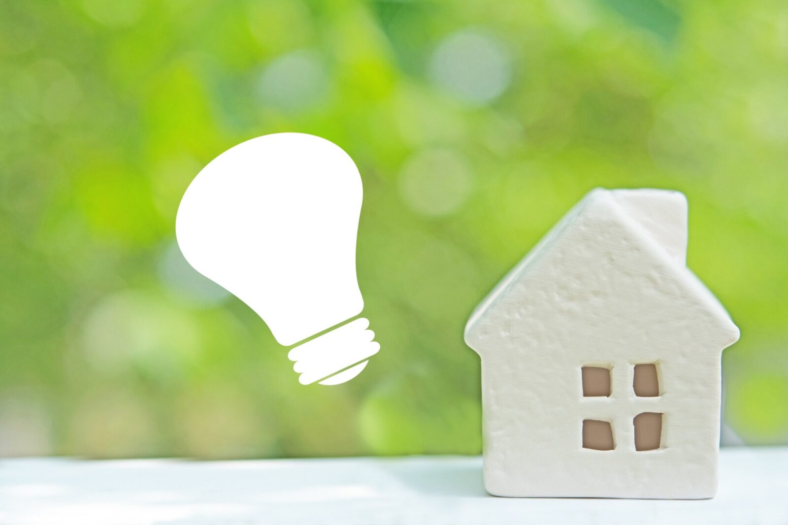 緑の背景に白い電球のアイコンとミニチュアの家。省エネ住宅やエコな暮らしのイメージ。