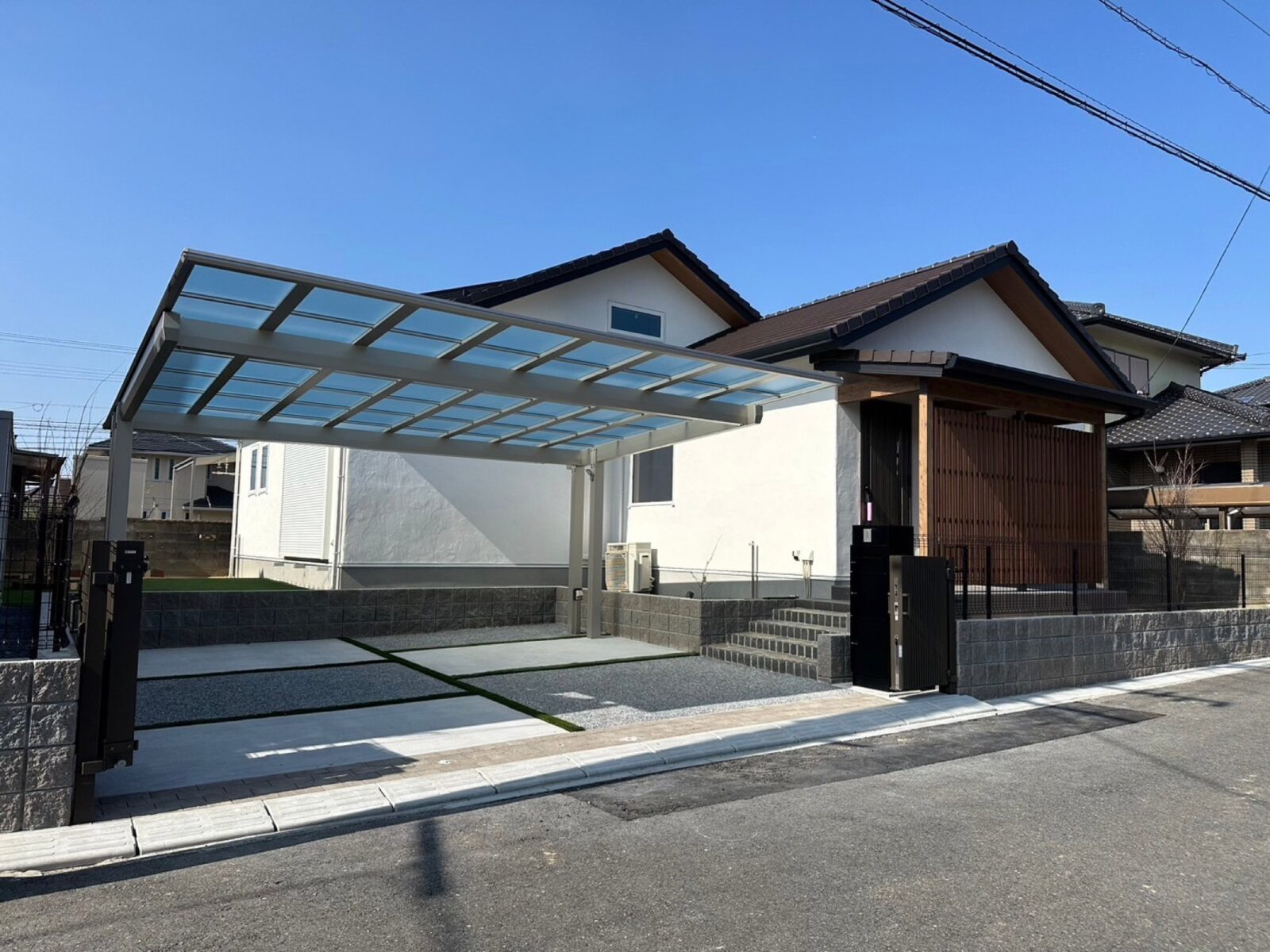 モダンな白い戸建て住宅の施工事例。透明なカーポートとデザインされた駐車スペース、木目調ルーバーの玄関アプローチが特徴の、機能的で統一感のある外構デザイン。