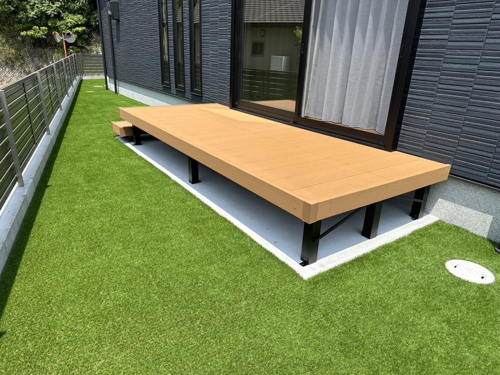 住宅の掃き出し窓の前に設置された一段ステップ付きの明るい木目調ウッドデッキと、その周囲に広がる鮮やかな緑の人工芝の庭。手入れが簡単で一年中美しいアウトドアリビング空間を実現。