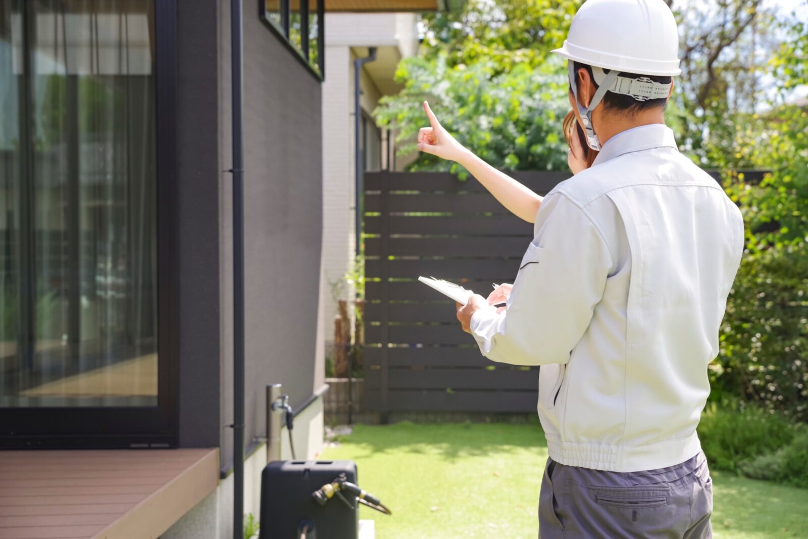 白いヘルメットと作業着の男性が、モダンな住宅の庭で書類に記入しながら女性の質問に答えている。新築住宅の点検、リフォーム、建築検査を行う専門家。