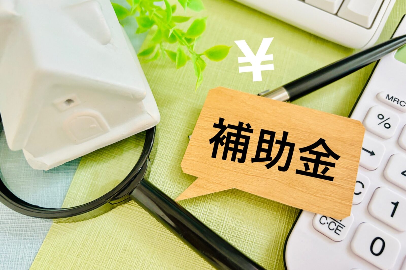 白い家の模型、虫眼鏡、電卓、ペン、¥マークが配置され、「補助金」と書かれた吹き出しボードが手前にある。住宅購入やリフォーム、事業支援などの補助金制度の検討、情報収集、資金計画を象徴する。