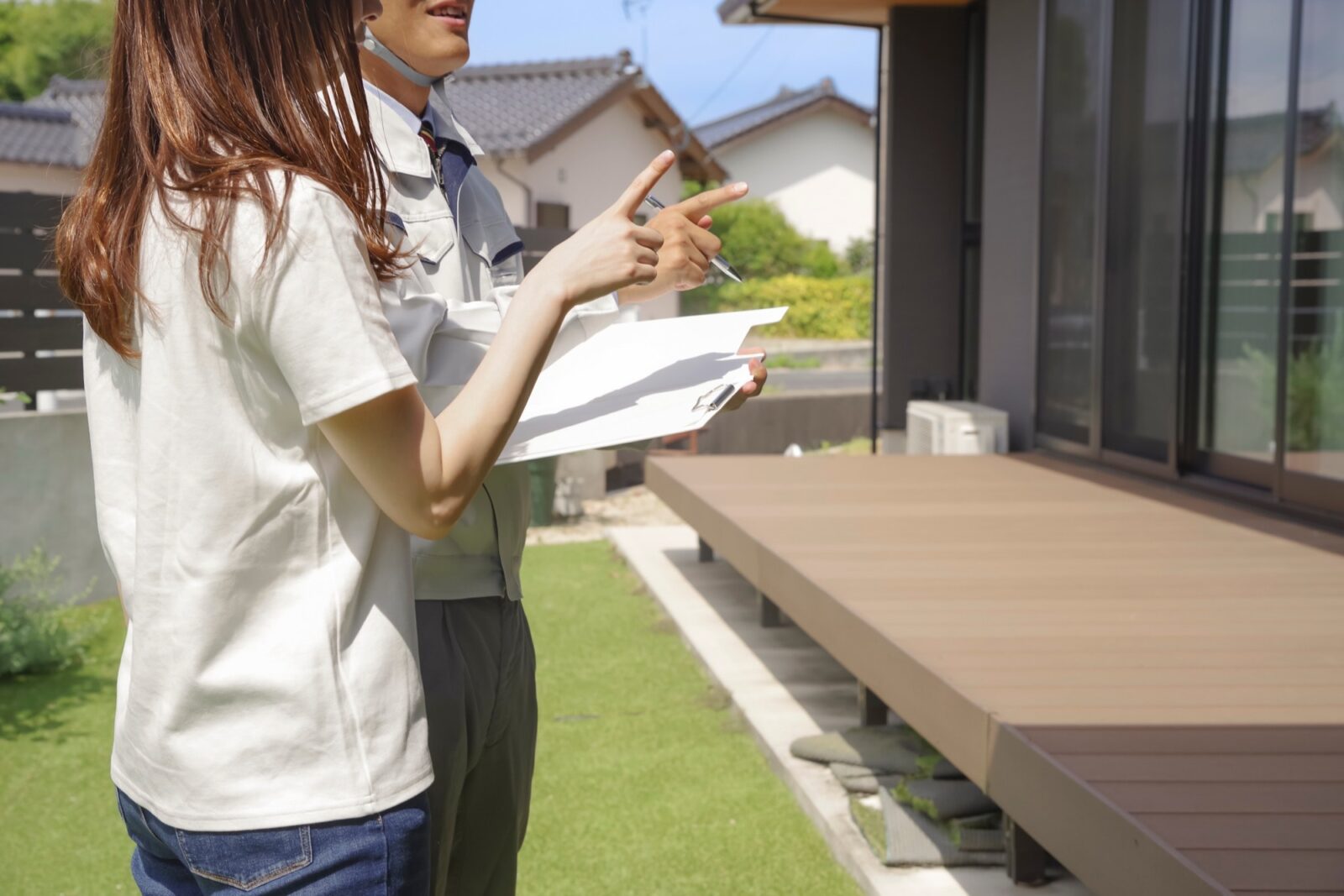 新築住宅の庭で、作業着の男性が女性にウッドデッキや家について指をさしながら説明している。専門家による住宅相談やリフォームの打ち合わせ風景。