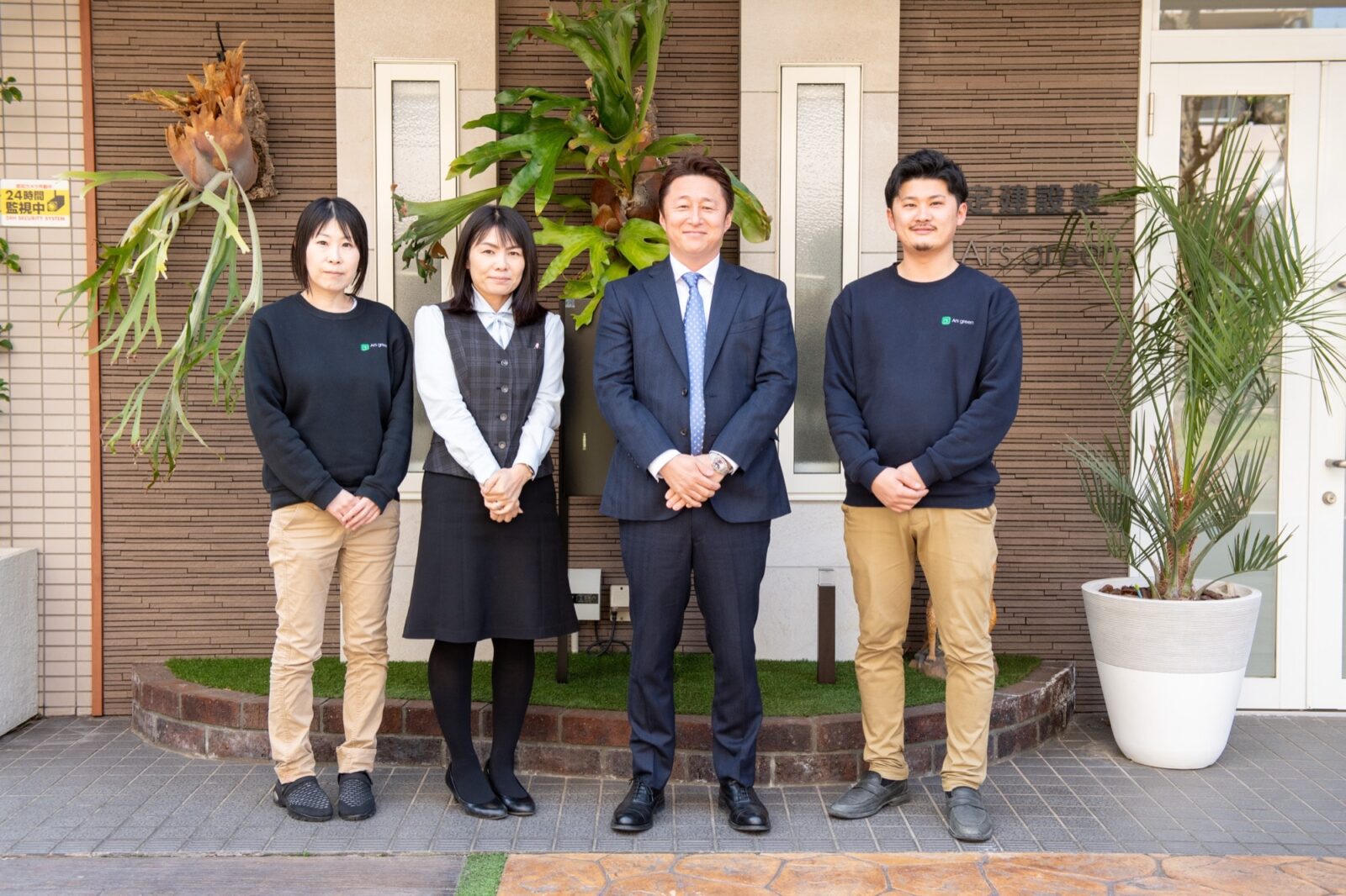 アルスグリーン株式会社のスタッフ4人が、会社の入り口前で笑顔で並ぶ。スーツ姿の男性代表、制服の女性社員、カジュアルな服装の男女社員が写り、背景には観葉植物が飾られている。